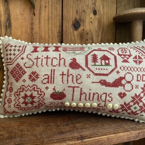 May include: A beige cushion with a white trim and red embroidery. The embroidery features a variety of designs, including a house, trees, birds, and the text "Stitch all the things".