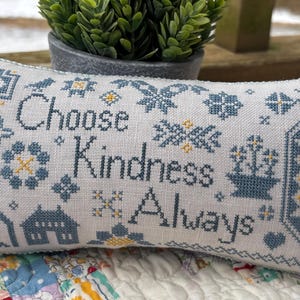 May include: A white pillow with blue and yellow embroidery. The pillow features the text "Choose Kindness Always" surrounded by various floral and geometric patterns.
