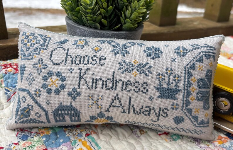 May include: A white pillow with blue and yellow embroidery. The pillow features the text "Choose Kindness Always" surrounded by various floral and geometric patterns.