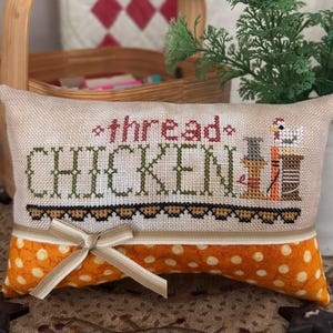 Pode incluir: Uma almofada decorativa com as palavras "thread CHICKEN" bordadas em vermelho e verde. O design inclui uma galinha e carretéis de linha. A seção inferior é laranja com bolinhas brancas e um laço bege.
