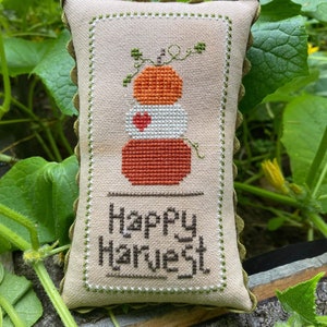 Puede incluir: Una almohada rectangular de tela beige con un diseño de punto de cruz de tres calabazas apiladas en tonos naranja y marrón, rematadas con vides verdes. Las palabras "Happy Harvest" están cosidas debajo. La almohada tiene un borde verde festoneado.