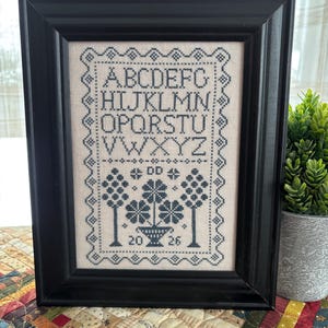 Puede incluir: Muestra de punto de cruz enmarcada en un marco negro. La muestra presenta el alfabeto, motivos florales y los números 20 y 26. El punto de cruz es de color azul oscuro sobre un fondo beige claro.