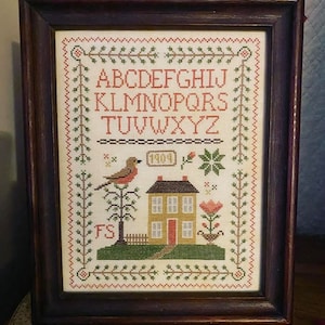 May include: A framed cross-stitch sampler featuring a house, a bird, and the alphabet. The house is yellow with a red door and a brown roof. The bird is red with a yellow breast. The alphabet is in red letters. The sampler is framed in a dark wood frame.