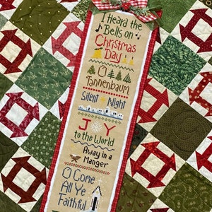 Puede incluir: Un sampler de punto de cruz beige con ribete rojo y blanco. El sampler presenta una variedad de textos navideños, incluyendo "I Heard the Bells on Christmas Day", "Tannenbaum", "Silent Night", "Joy to the World", "Away in a Manger", y "O Come All Ye Faithful".