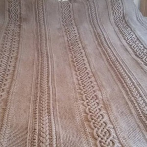 May include: A beige knitted throw blanket with a cable knit pattern. The blanket is folded and laid out on a bed.