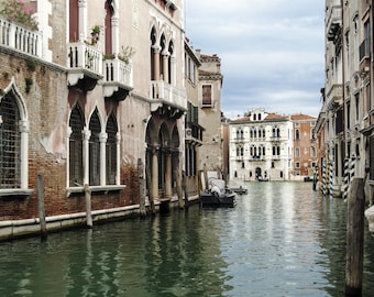 May include: A narrow canal in Venice, Italy, with buildings lining both sides. The buildings are old and have a rustic, weathered look. The water is calm and reflects the sky and buildings. A small boat is in the water.
