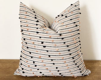May include: Square cushion with a cream-coloured background, black and copper-coloured horizontal stripes, and small, raised black and copper-coloured dots. The cushion is on a wooden surface.
