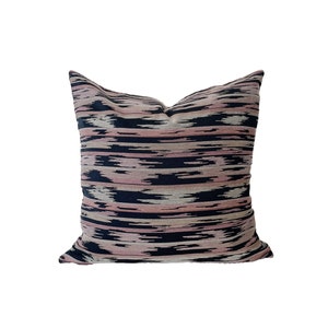 May include: Square decorative pillow with a horizontal ikat pattern in shades of pink, navy blue, and beige. The pillow has a soft, textured appearance and is likely made of fabric. The pillow is designed for home decor.