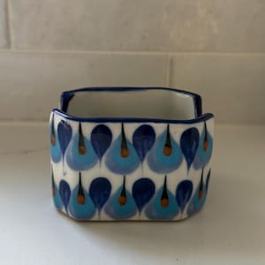 May include: A square ceramic napkin holder featuring a white base with a repeating pattern of blue and light blue teardrop shapes, each accented with a small brown center. The rim of the holder is outlined in a dark blue glaze.