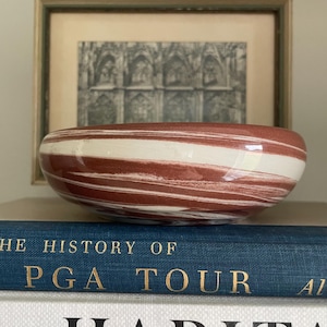 Puede incluir: Un cuenco decorativo ovalado con un diseño en espiral rojo y blanco. El cuenco de cerámica está sobre una pila de libros, con una imagen enmarcada en el fondo. Un elemento decorativo único.