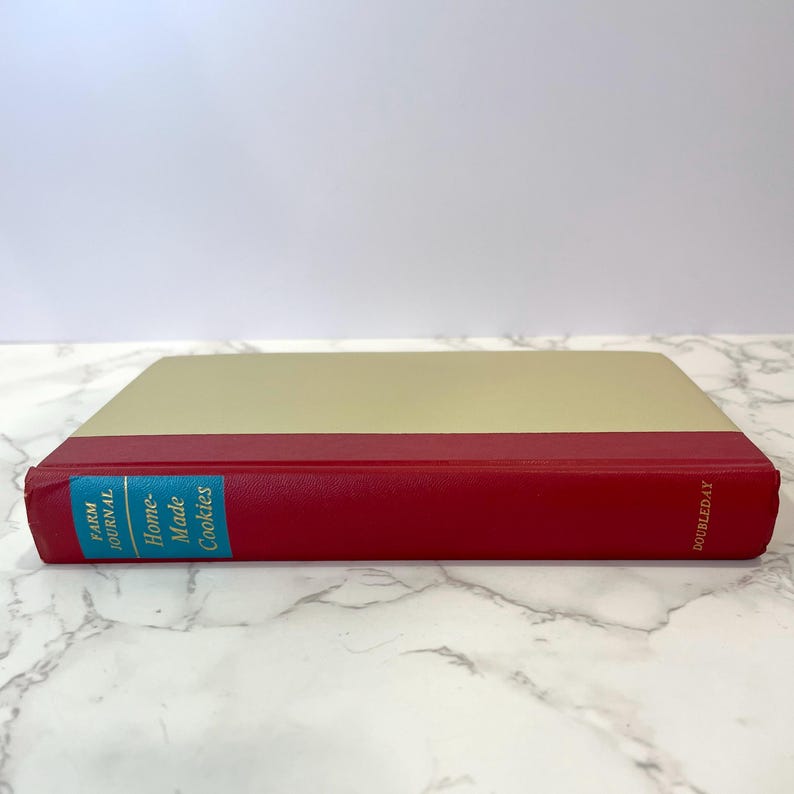 K&ouml;nnte beinhalten: Ein rotes Hardcover-Buch mit einem cremefarbenen R&uuml;cken. Der Buchtitel lautet "Home Made Cookies" in blauer Schrift auf t&uuml;rkisfarbenem Hintergrund. Das Buch stammt aus der Reihe "Farm Journal" und wird von Doubleday herausgegeben.