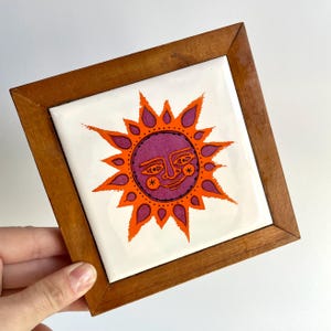 May include: A square ceramic tile set in a brown wooden frame. The tile features a white background with a central graphic of a sun with a purple face, orange rays, and black line details. The sun has a stylized expression with circular cheeks.