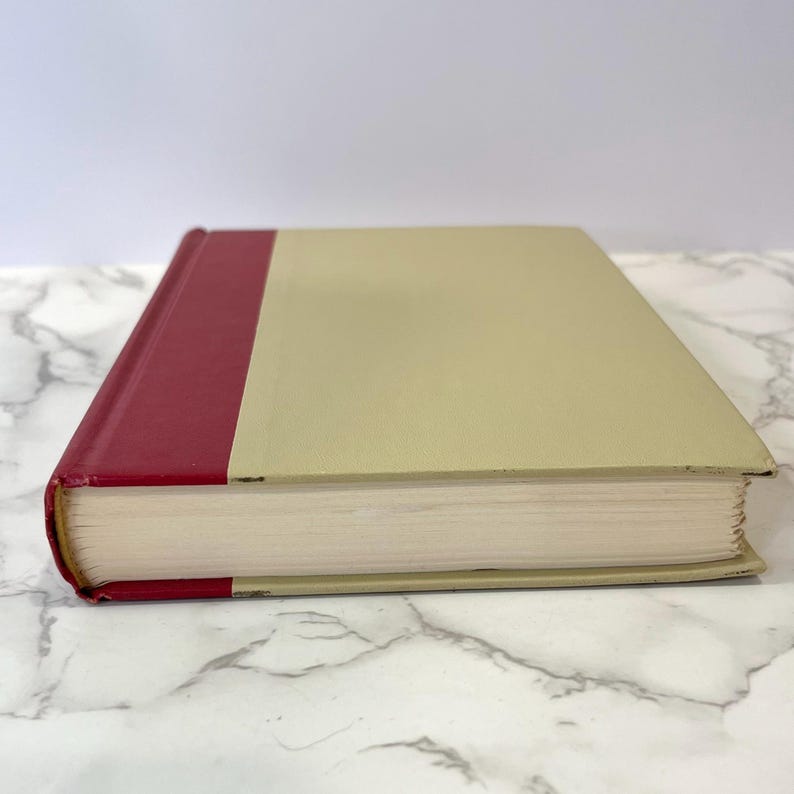 K&ouml;nnte beinhalten: Ein gebundenes Buch mit einem cremefarbenen Einband und einem roten R&uuml;cken. Das Buch ist geschlossen und liegt auf einer wei&szlig;en Marmorfl&auml;che.