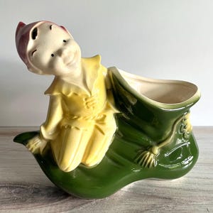 May include: A ceramic planter shaped like a green shoe with a yellow-clad elf figure attached to the side. The elf has a pink hat, a smiling face, and is positioned as if leaning against the shoe. The interior of the shoe is cream-colored.