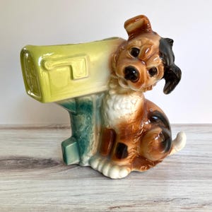 May include: A ceramic dog planter with a mailbox design. The dog is brown, black, and white, with large eyes. The mailbox is light green and blue, adding a pop of colour to the vintage decor. A charming piece for any home.
