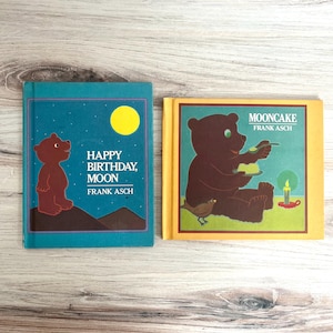 Pode incluir: Dois livros infantis com ilustrações de ursos. Um livro tem uma capa azul-turquesa com um urso castanho e o texto "Happy Birthday, Moon" de Frank Asch. O outro livro tem uma capa amarela com um urso castanho comendo bolo e o texto "Mooncake" de Frank Asch.