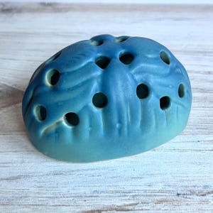 May include: A blue ceramic flower frog with multiple holes for arranging stems. The frog has a rounded shape and a textured surface, with a gradient of blue hues. It is designed to hold flowers in place.