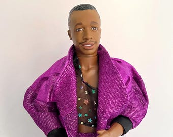May include: A doll wearing a shiny purple suit with black trim. The doll has a black mesh top with colorful star accents. The doll has a dark complexion and a smiling expression.