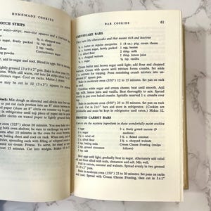 K&ouml;nnte beinhalten: Ein altes Kochbuch, das auf einer Seite mit Rezepten f&uuml;r K&auml;sekuchen-Riegel und gefrostete Karotten-Riegel ge&ouml;ffnet ist. Die Rezepte beinhalten Zutaten wie Butter, Zucker, Mehl, Eier, Frischk&auml;se, Karotten und Kokosnuss.