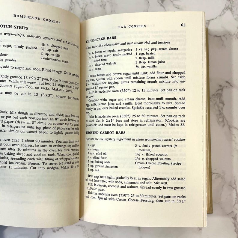 K&ouml;nnte beinhalten: Ein altes Kochbuch, das auf einer Seite mit Rezepten f&uuml;r K&auml;sekuchen-Riegel und gefrostete Karotten-Riegel ge&ouml;ffnet ist. Die Rezepte beinhalten Zutaten wie Butter, Zucker, Mehl, Eier, Frischk&auml;se, Karotten und Kokosnuss.