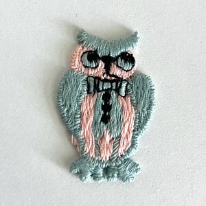 Könnte beinhalten: Gestickter Eulenaufnäher in Blau- und Rosatönen. Die Eule hat große schwarze Augen und eine Fliege. Der Aufnäher ist wahrscheinlich für Kleidung oder Accessoires gedacht.