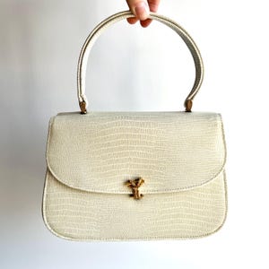 May include: Cream-colored handbag with a textured, reptile-like pattern. The bag features a curved handle and a gold-colored clasp. The overall design is classic and elegant, suitable for various occasions.