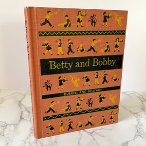 May include: A vintage children's book titled "Betty and Bobby" by Durrell and Sullivan. The cover features a pink background with black and yellow illustrations of children playing.