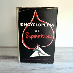 Könnte beinhalten: Ein Vintage-Buch mit dem Titel "Encyclopedia of Superstitions" mit einem schwarzen Einband mit einem roten und weißen Pik-Design. Der Titel ist in Weiß und das Wort "Superstitions" in Rot. Das Buch steht aufrecht.