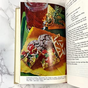 K&ouml;nnte beinhalten: Offenes Kochbuch mit einem Rezept f&uuml;r Festtagskekse. Das Buch zeigt ein Foto der Kekse, darunter Chocolate Cracklins, Toffee Sticks und Fruitcake Squares. Der Text lautet "Celebration Cookies. Dekorativ, aber einfach zu machen: Chocolate Cracklins, oben (Seite 192); Toffee Sticks, rechts (Seite 66); Fruitcake Squares (Seite 236). Sie sehen - und schmecken - schick aus."
