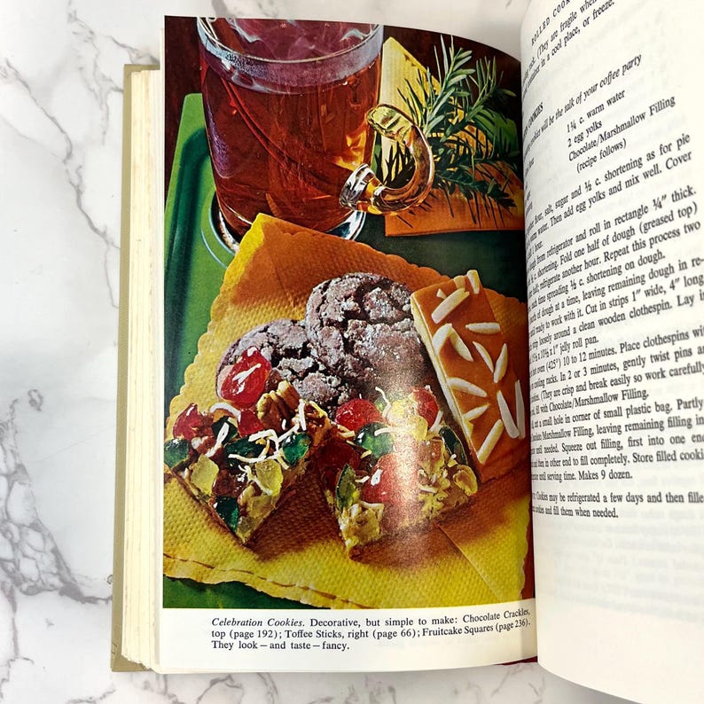 K&ouml;nnte beinhalten: Offenes Kochbuch mit einem Rezept f&uuml;r Festtagskekse. Das Buch zeigt ein Foto der Kekse, darunter Chocolate Cracklins, Toffee Sticks und Fruitcake Squares. Der Text lautet "Celebration Cookies. Dekorativ, aber einfach zu machen: Chocolate Cracklins, oben (Seite 192); Toffee Sticks, rechts (Seite 66); Fruitcake Squares (Seite 236). Sie sehen - und schmecken - schick aus."