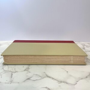 K&ouml;nnte beinhalten: Ein geschlossenes Buch mit einem beigefarbenen Einband und einem d&uuml;nnen roten Band oben.
