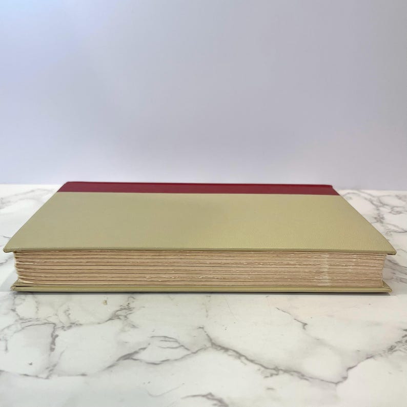 K&ouml;nnte beinhalten: Ein geschlossenes Buch mit einem beigefarbenen Einband und einem d&uuml;nnen roten Band oben.