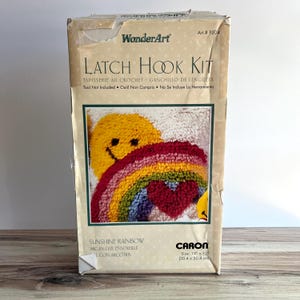 May include: A WonderArt Latch Hook Kit in a rectangular cardboard box. The packaging features a graphic of a yellow smiley face, a colourful rainbow, and a red heart. Text includes Sunshine Rainbow and a size of 30.4 cm by 30.4 cm.
