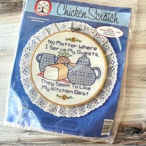 May include: A packaged embroidery kit featuring a circular design with the text "No Matter Where I Serve My Guests, They Seem To Like My Kitchen Best." The design includes embroidered images of a teapot, bread, and pineapple. The kit includes a wooden hoop and stamped design.