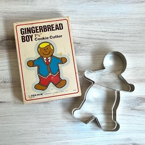 May include: A vintage gingerbread boy cookie cutter and its original box. The box features a cartoon gingerbread boy in a blue jacket, red pants, and a yellow hat. The cutter is 7.5 inches.
