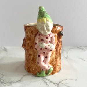 May include: A ceramic planter shaped like a tree trunk, with a figurine of a gnome in pink pyjamas with red polka dots, a green hat, and green boots. The planter has a brown, textured exterior.