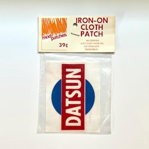 May include: A rectangular iron-on cloth patch in a clear plastic bag with a header card. The patch features a blue circle behind a vertical red rectangle with the white text DATSUN. The header card reads IRON-ON CLOTH PATCH and mod patches.