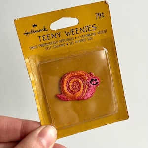 May include: A vintage Hallmark "Teeny Weenies" self-sticking Swiss embroidered applique. The applique is a smiling snail with a pink body and an orange and yellow shell. The packaging is yellow with text.
