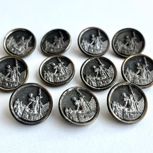 May include: A set of antique silver-coloured buttons. Each button displays a detailed relief of a figure with a spear, set against a backdrop of a fortress or wall. The buttons are round and have a slightly aged appearance.
