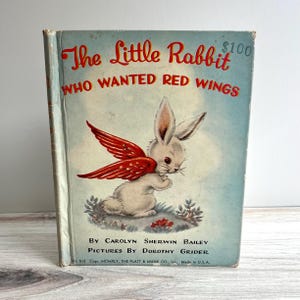 Puede incluir: Libro infantil vintage titulado "The Little Rabbit Who Wanted Red Wings". La portada presenta un conejo blanco con alas rojas sobre un fondo azul claro. El título está en rojo, con los nombres del autor y el ilustrador debajo. Hecho en EE. UU.
