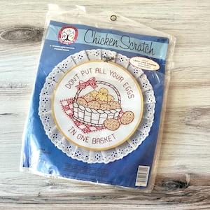 May include: A Chicken Scratch embroidery kit in a plastic package. The kit features a round design with the text "DON'T PUT ALL YOUR EGGS IN ONE BASKET" surrounding a basket of eggs and a chick. The design is framed by a white lace-like border.