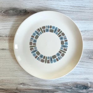 May include: A cream-colored ceramic plate with a circular design of geometric shapes in blue, brown, and black. The plate has a wide rim and a central area free of decoration. The design is a repeating pattern of rectangles, ovals, and starbursts.