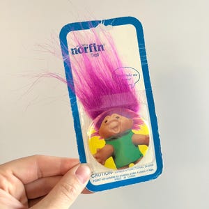 May include: A vintage Norfin Troll doll in its original packaging. The doll has bright pink hair and a smiling face. The packaging is blue and white with the text "Adopt a Norfin Troll". The doll is a collectible toy.