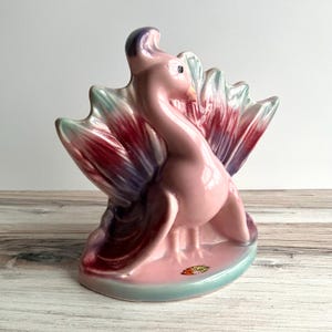May include: A vintage ceramic flamingo figurine in shades of pink, red, purple, and teal. The flamingo has a long neck and is standing in front of a fan-shaped design. The figurine is a decorative item.