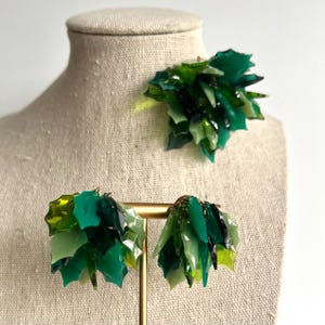 May include: A set of green earrings and a matching brooch. The jewelry features a cluster of irregularly shaped, translucent green pieces in varying shades, creating a layered, textured effect. The earrings are suspended from gold-colored posts, and the brooch is pinned to a mannequin.
