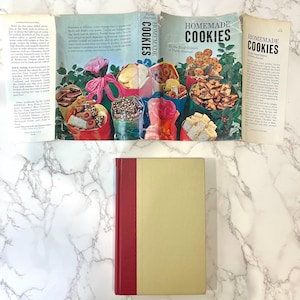 K&ouml;nnte beinhalten: Ein Vintage-Kochbuch mit dem Titel "Homemade Cookies" von den Food Editors des Farm Journal. Der Umschlag zeigt eine farbenfrohe Illustration verschiedener Kekse in dekorativen Papierkegeln. Das Buch ist geschlossen und liegt auf einer wei&szlig;en Marmorfl&auml;che.