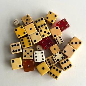May include: A collection of vintage dice in various colors, including ivory, yellow, and red. The dice have black dots indicating the numbers, and are scattered across a white surface. The dice are of different sizes and materials.