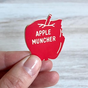 May include: A red apple-shaped pin with the words "APPLE MUNCHER" in white. The apple has a bite taken out of it, and the pin is held by a hand.