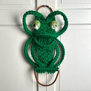 May include: Green macrame wall hanging in the shape of a frog with large, white eyes and a wooden ring at the top.
