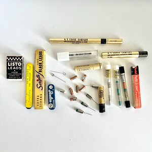 May include: An assortment of vintage writing supplies, including pencil lead boxes, erasers, and mechanical pencils. The items are in various colors, including black, gold, yellow, and white. The image features several brands, such as Scripto and Cross.